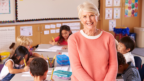 Teacher smilining and in the background a group of children sitting on a table doing an assignment.