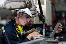 A man wearing a hardhat next to machinery, using a laptop